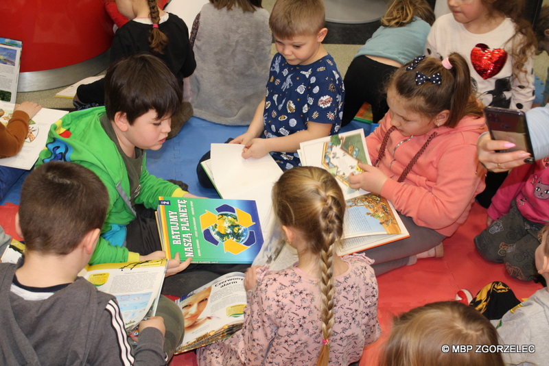 W pomieszczeniu biblioteki, grupa przedszkolaków siedzi na podłodze i przegląda książki o tematyce ekologicznej.