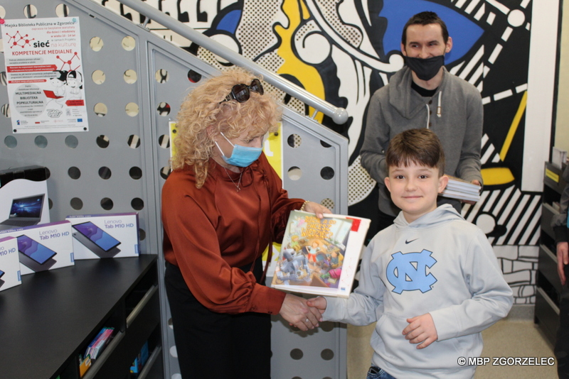 W pomieszczeniu biblioteki Dyrekcja MBP w Zgorzelcu wręcza chłopcu nagrodę książkową i zaświadczenie udziału w projekcie "Sieć na kulturę". Za nimi stoi bibliotekarz.