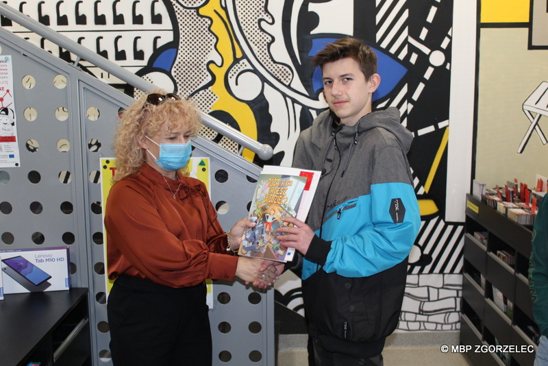 W pomieszczeniu biblioteki Dyrekcja MBP w Zgorzelcu wręcza chłopcu nagrodę książkową i zaświadczenie udziału w projekcie "Sieć na kulturę". Za nimi stoi bibliotekarz.
