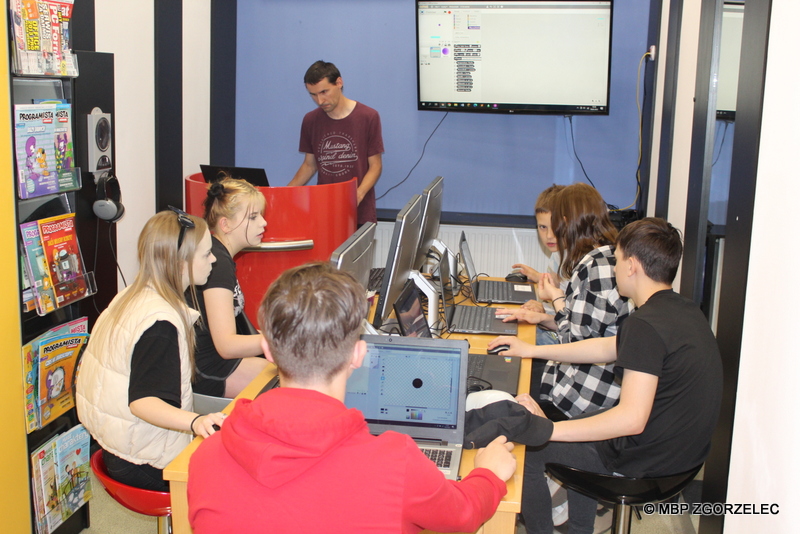 W pomieszczeniu Mediateki jest widocznych 6 osób pracujących przy komputerach. Z tyłu stoi bibliotekarz i wyjaśnia na ekranie telewizora funkcje obsługi programu Scratch
