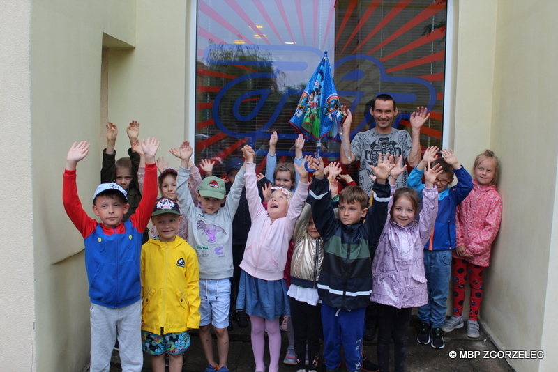 Grupa przedszkolaków z bibliotekarzem, stoi pod oknem Mediateki z uniesionymi rękoma do góry