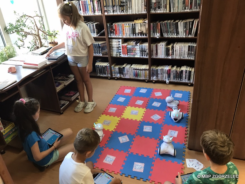 W pomieszczeniu biblioteki po piankowej macie jeżdżą zaprogramowane roboty. Obok siedzą dzieci i odkrywają wybrane pary kart w grze memory.