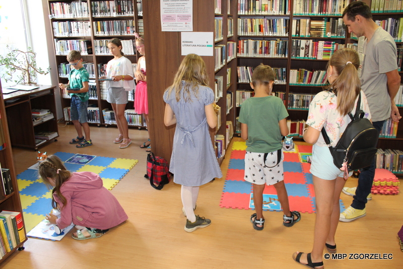 W pomieszczeniu biblioteki jest 8 osób. Dzieci programują roboty na piankowej macie a obok stoi bibliotekarz i przygląda się ich pracy.