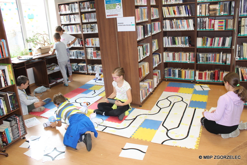W pomieszczeniu biblioteki po piankowej macie jeżdżą zaprogramowane roboty. Obok siedzą dzieci i programują trasy dla robotów z użyciem białych arkuszy z nadrukowaną czarną linią.