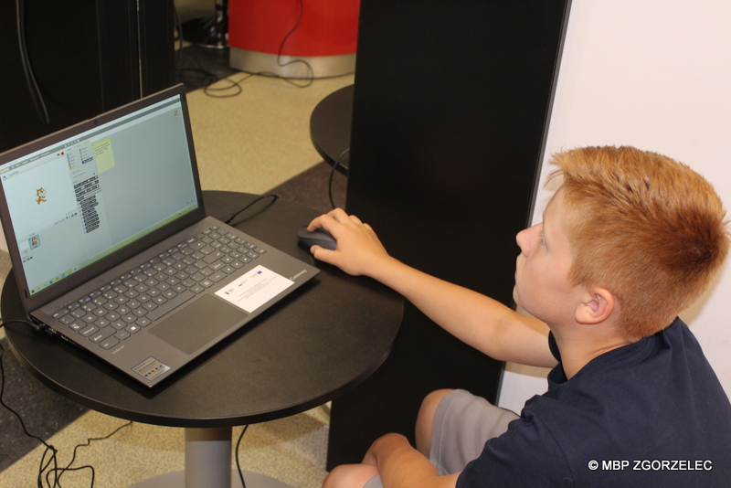 W pomieszczeniu biblioteki siedzi chłopiec przy komputerze i programuje grę.