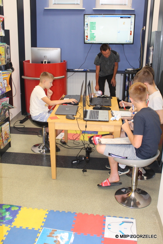 W pomieszczeniu biblioteki bibliotekarz schyla się do podłogi. Obok przy stolikach siedzi czterech chłopców. Na komputerach programują roboty.