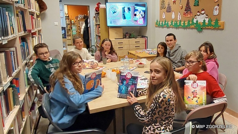 W pomieszczeniu czytelni Oddziału Dla Dzieci i Młodzieży MBP w Zgorzelcu siedzi przy stołach 9 osób. Kilka dziewczyn trzyma w rękach czasopismo młodzieżowe.