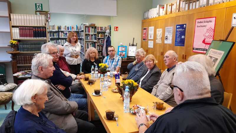 Grupa osób siedzi i rozmawia przy stole w bibliotecznej czytelni. Na stole znajdują się filiżanki kawy, butelki z wodą i kwiaty.
