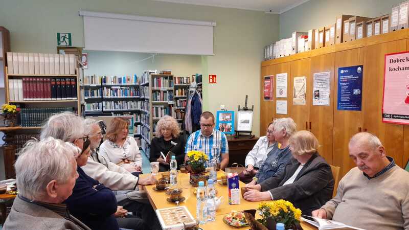 Grupa osób siedzi i rozmawia przy stole w bibliotecznej czytelni. Na stole znajdują się filiżanki kawy, butelki z wodą i kwiaty.