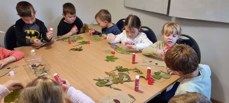 Grupa dzieci wykonuje prace plastyczne, siedząc przy stole. Na stole leżą liście i kleje w sztyfcie.