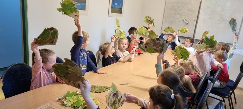 Grupa dzieci siedzi przy stole, chwaląc się wykonanymi przez siebie pracami plastycznymi, unosząc je nad głowy.