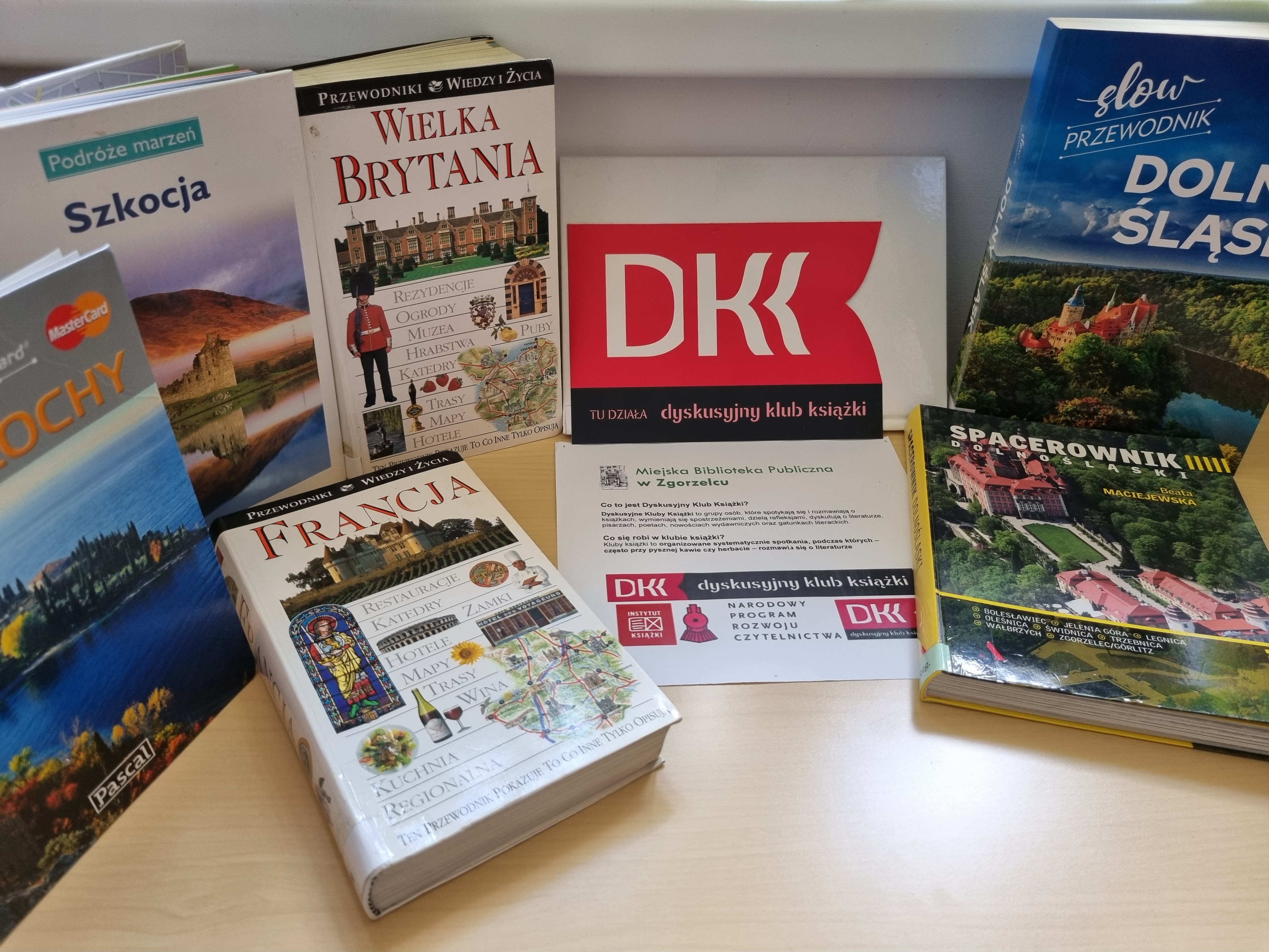 Na stole z jasnego drewna leżą książki podróżnicze i przewodniki po różnych krajach oraz kartka z informacją o klubie DKK.