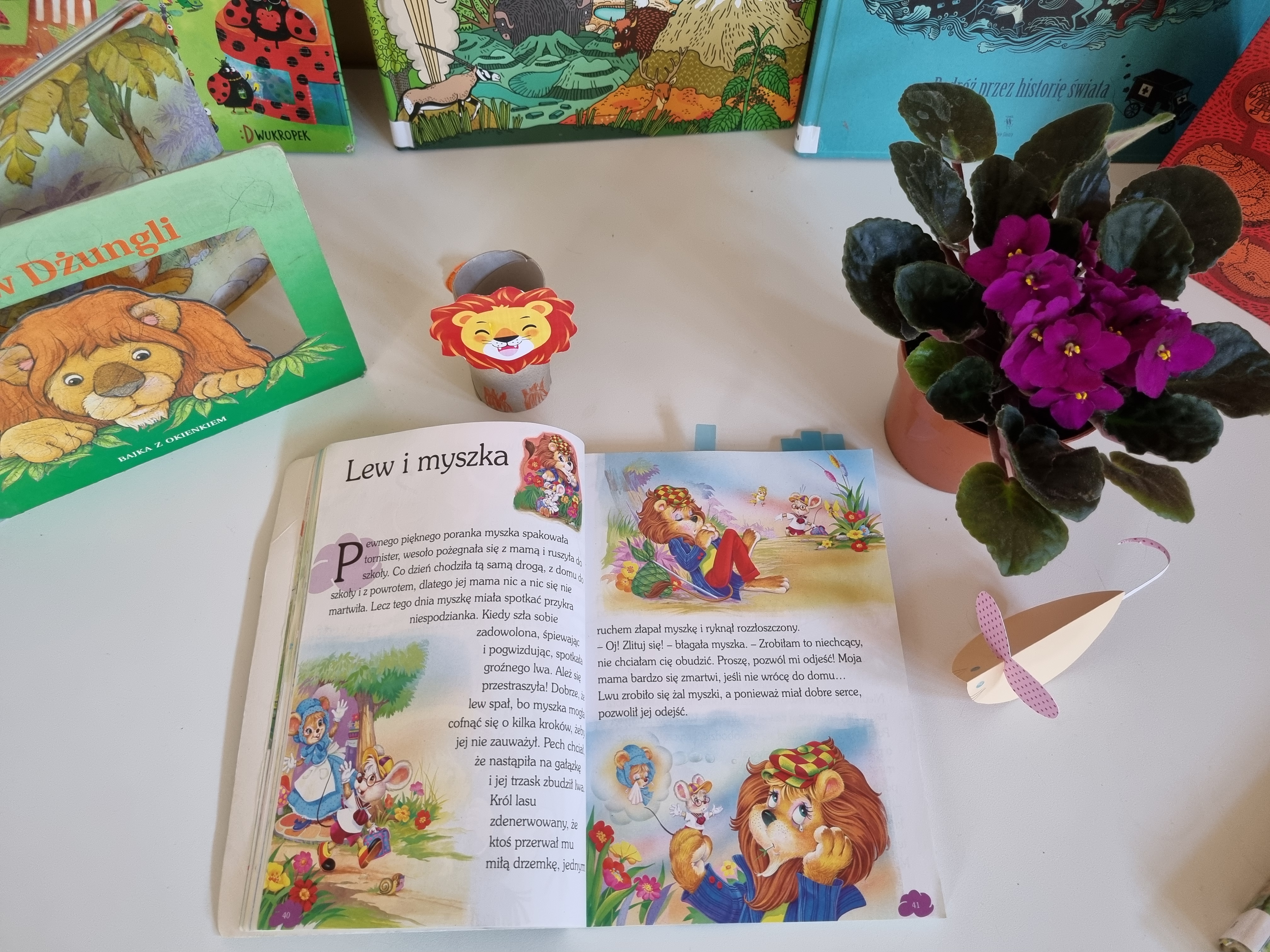 Na białym stole leży otwarta książka z opowiadaniem zatytułowanym "Lew i myszka". Wokół znajdują się inne książki oraz ozdoby z papieru przedstawiające lwy.