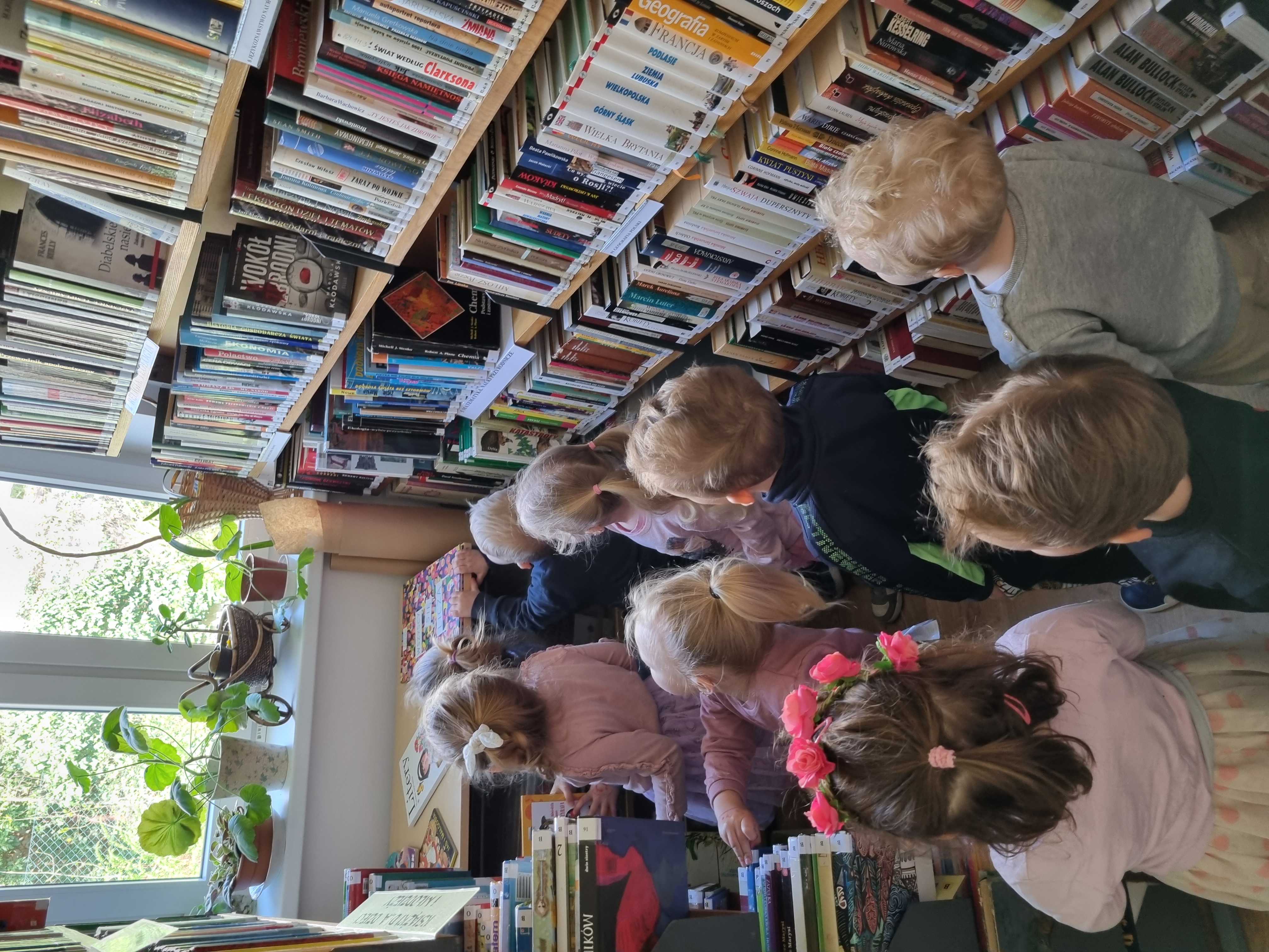 Grupa dzieci w wieku przedszkolnym przygląda się książkom w bibliotece, stojąc obok regału.