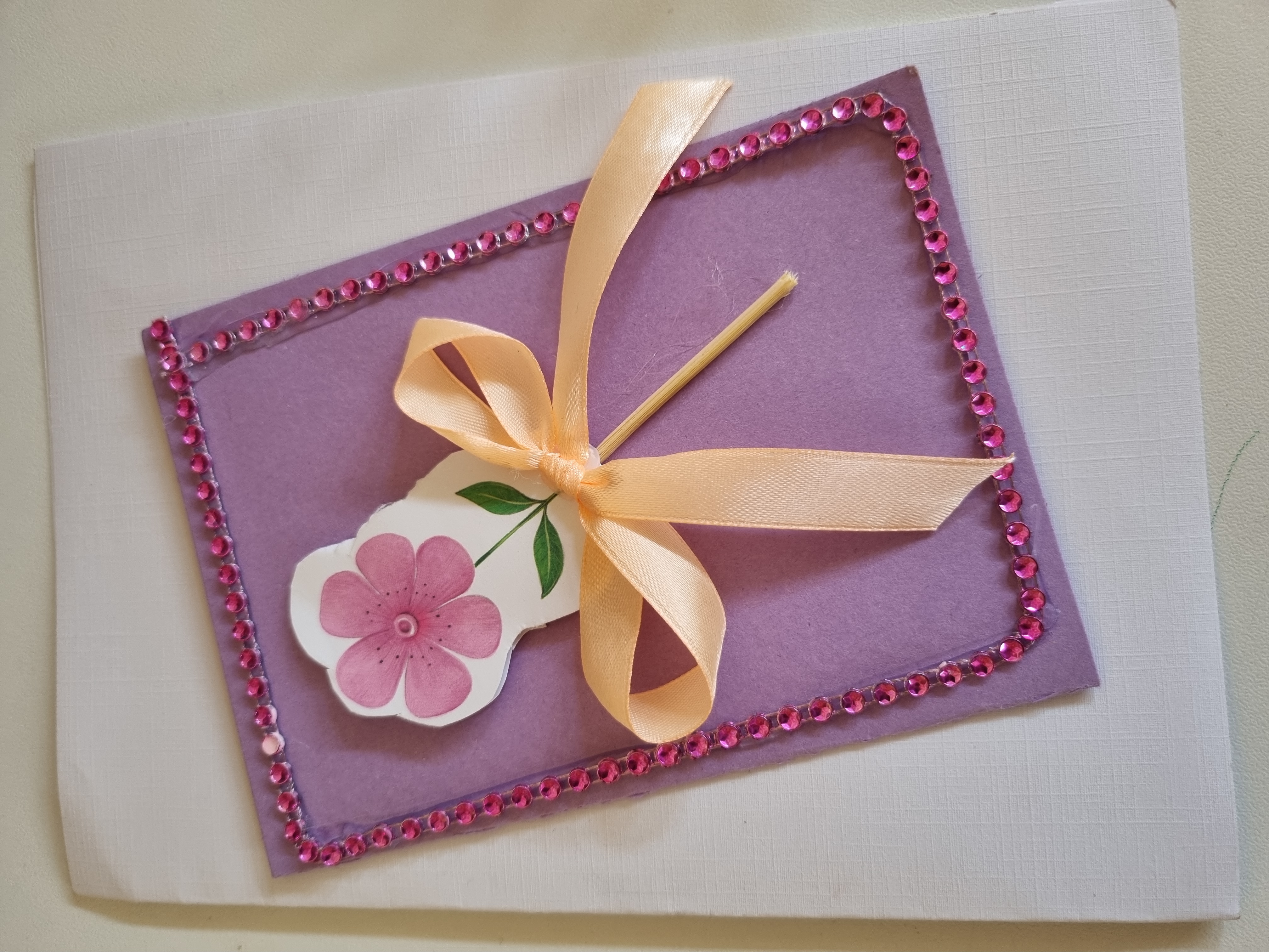 Na jasnym tle znajduje się praca plastyczna z papieru i innych materiałów, przedstawiajaca fioletowy kwiat, obwiązany kokardą.