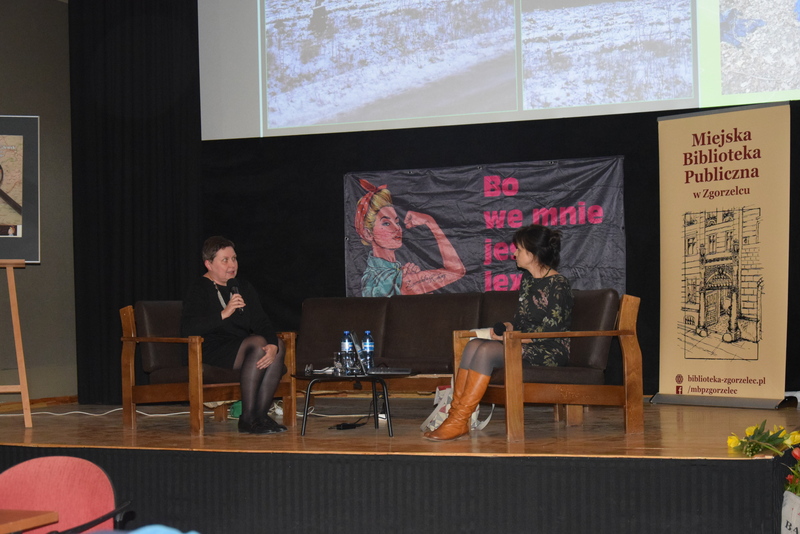 Dwie kobiety siedzą na niskich, obitych czarną skórą fotelach pośrodku sceny, rozmawiając przez mikrofony. W tle znajduje się slajd prezentacji i banery organizatorów spotkania.