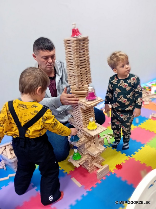 W pomieszczeniu hali sportowej, przy stanowisku Miejskiej Biblioteki Publicznej w Zgorzelcu, trzy osoby budują wieżę z drewnianych klocków.