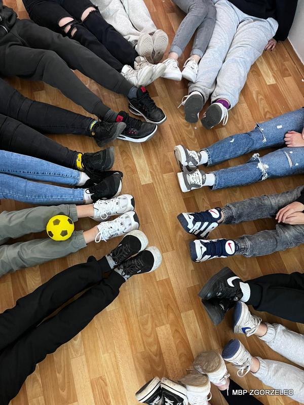 Zdjęcie przedstawiające widok z góry nóg uczestników zajęć siedzących na podłodze. Na kolanach jednego z uczestników jest piłka.