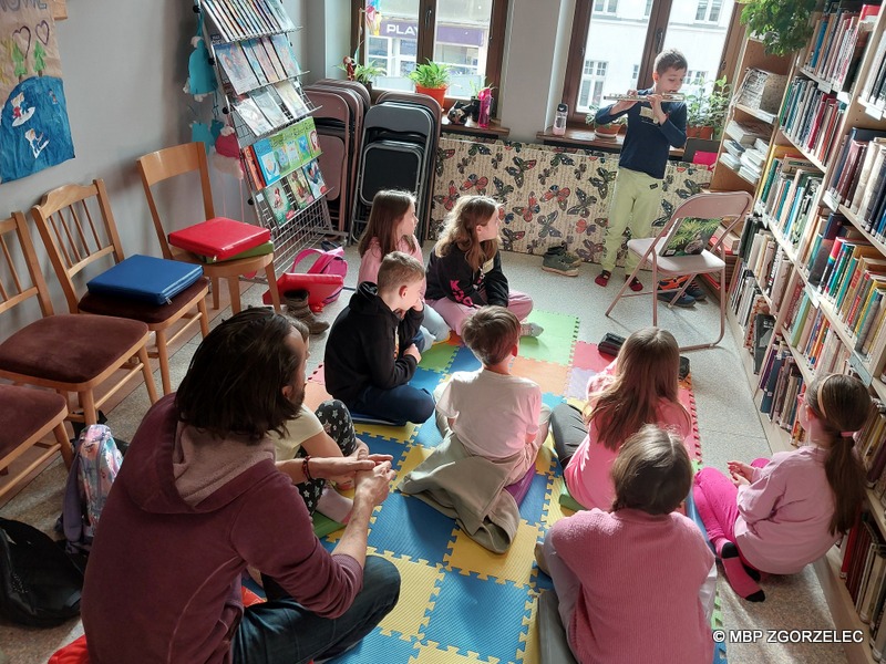 W pomieszczeniu Oddziału Dla Dzieci Miejskiej Biblioteki Publicznej w Zgorzelcu, grupa dzieci słucha umiejętności muzyczne jednego z uczestników zajęć.