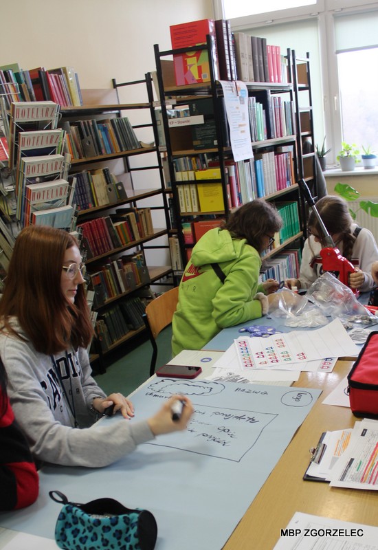 W pomieszczeniu biblioteki szkolnej uczniowie projektują odręcznie plakat i tworzą przypinki promocyjne projektu "Książki w chmurach"