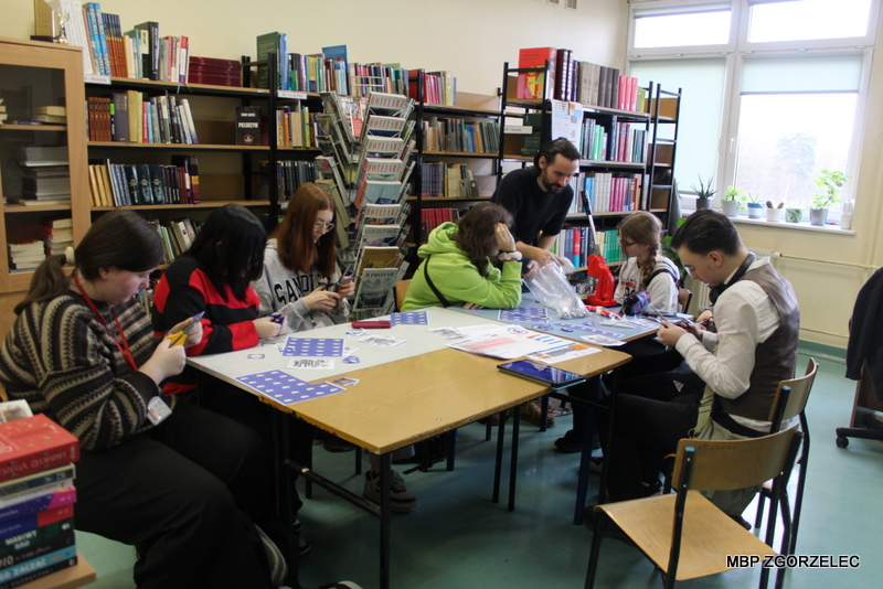 W pomieszczeniu biblioteki szkolnej uczniowie tworzą przypinki promocyjne projektu "Książki w chmurach"