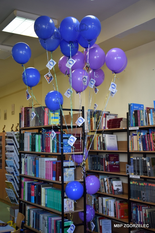 W pomieszczeniu biblioteki, na tle regałów z książkami', unoszą się niebieskie i fioletowe balony z logiem kampanii "Książki w chmurach" wraz z kodami dostępowymi do e-podręczników