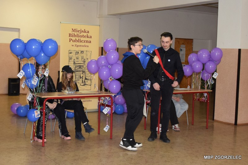 Na korytarzu szkolnym jest sześć młodych osób w otoczeniu niebieskich i fioletowych balonów. Dwoje młodych uczniów na zmianę przemawiają do mikrofonu.