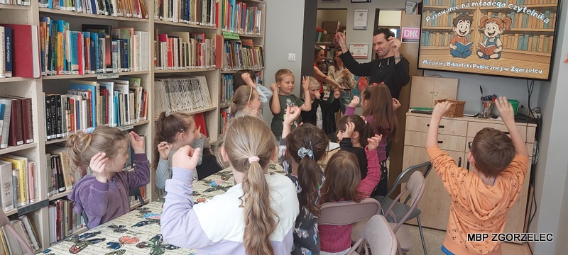 W pomieszczeniu biblioteki, dzieci wspólnie z bibliotekarzem unoszą ręce.