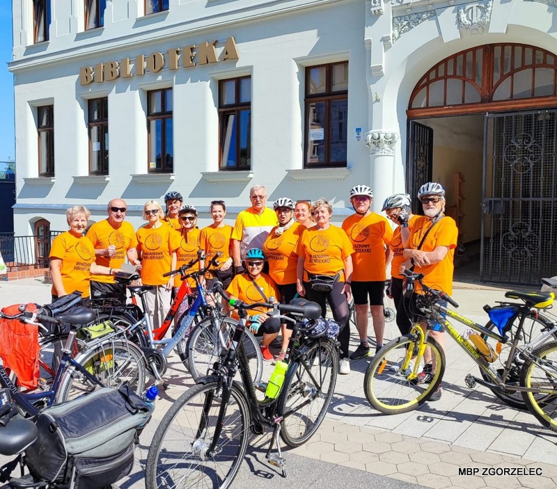 Przed budynkiem biblioteki stoją uczestnicy rajdu rowerowego
