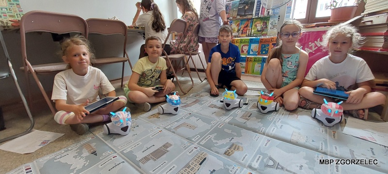 Dzieci sterujące robotami za pomocą tabletów na edukacyjnej macie z mapą w czytelni Oddziału Dla Dzieci i Młodzieży.