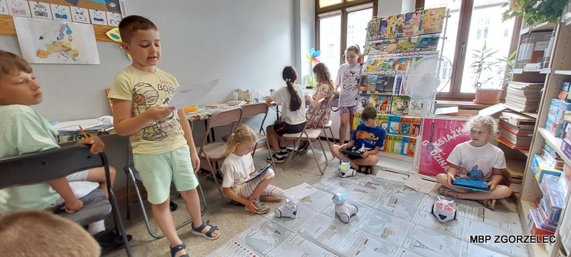 Dzieci sterujące robotami za pomocą tabletów na edukacyjnej macie z mapą w czytelni Oddziału Dla Dzieci i Młodzieży.