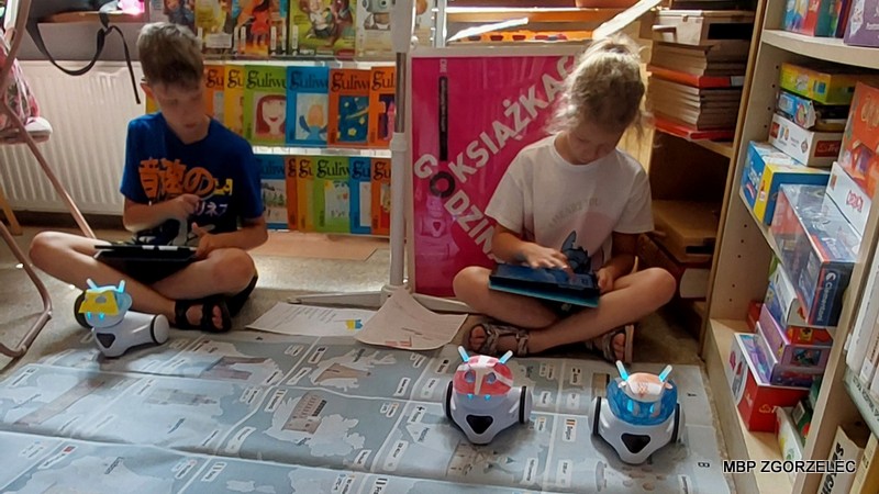Dzieci sterujące robotami za pomocą tabletów na edukacyjnej macie z mapą w czytelni Oddziału Dla Dzieci i Młodzieży.