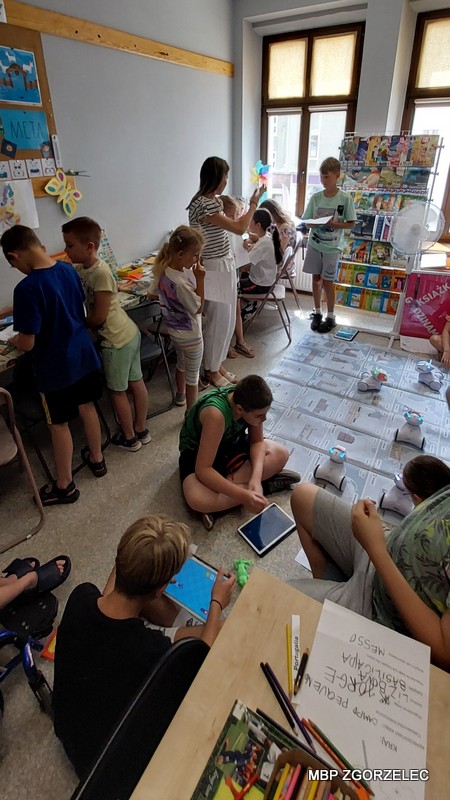Zdjęcie przedstawia czytelnię Oddziału Dla Dzieci i Młodzieży, w której dzieci oraz bibliotekarka uczestniczą w różnych aktywnościach. Część dzieci siedzi przy stołach, pracując nad pracami plastycznymi lub korzystając z tabletów, inne stoją lub siedzą na podłodze.