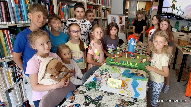 Grupa dzieci stojących wokół stołu w bibliotece. Na stole znajduje się kolorowa makieta z różnymi elementami, takimi jak budynki, drzewa i postacie. W tle widać półki z książkami oraz ekran z wyświetlonym obrazem fragmentu filmu Muminki..