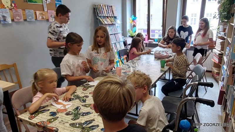 Grupa dzieci uczestniczących w zajęciach plastycznych w bibliotece. Dzieci siedzą wokół dwóch stołów, na których znajdują się różnorodne materiały artystyczne, takie jak farby, pędzle, klej i papier. W tle widoczne są regały z książkami.