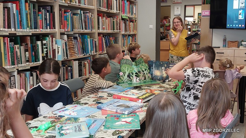 Grupa dzieci siedzących wokół stołu w bibliotece. Na stole znajdują się książki dla dzieci o Muminkach. W tle widoczne są regały pełne książek. Bibliotekarka stoi z przodu, trzymając książkę i czyta fragment dzieciom książki o Muminkach.