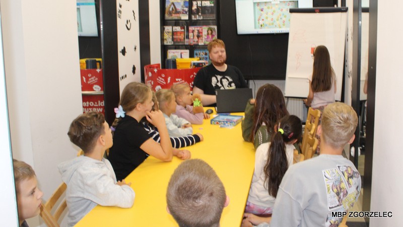 Grupa dzieci z bibliotekarzem przy żółtym stole w sali Mediateki grają w kalambury.