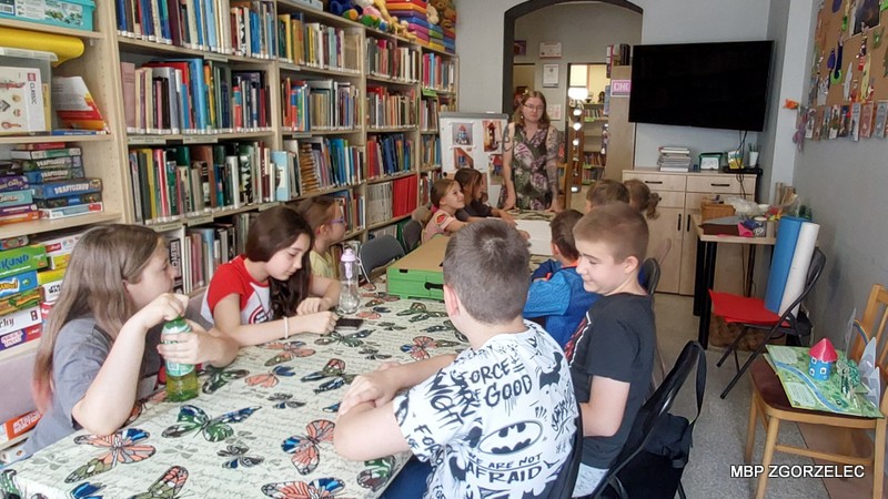 Dzieci siedzą przy stole w bibliotece i rozmawiają z bibliotekarką na temat zajęć.