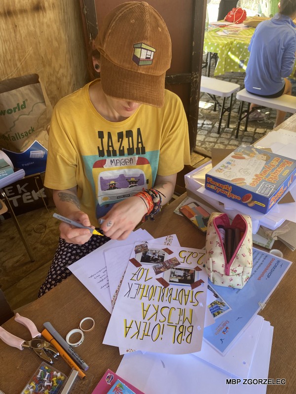 Bibliotekarka projektuje stronę w kronice Kindermiasta w czasie wydarzenia plenerowego. Na stole znajdują się markery, piórnik, taśma i pudełka. W tle widoczne są inne osoby przy stołach z zielonymi obrusami w kropki