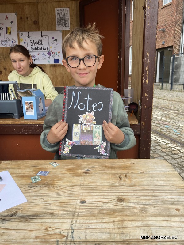 Chłopiec siedzący przy drewnianym stole trzyma w rękach ozdobiony zeszyt z napisem 'Notes'. W tle widoczne jest stoisko biblioteczne z dekoracjami i szyldem 'Stadt bibliothek', a na stole leżą drobne przedmioty.
