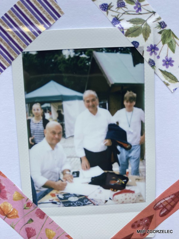 Fotografia przedstawia burmistrzów miast Zgorzelca i Gorlitz przy stole wypełniających karty do kroniki Kindermiasta. W tle widoczny jest namiot oraz zabudowania. Zdjęcie oprawione jest dekoracyjną taśmą z wzorami w paski, kwiaty i liście.
