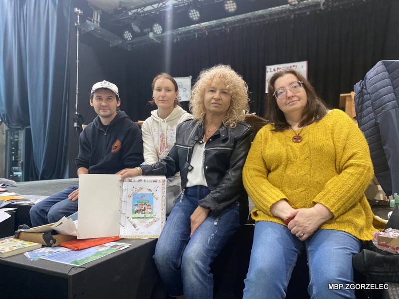 Zdjęcie przedstawia cztery osoby siedzące (bibliotekarzy z dyrekcją) na scenie, z których jedna trzyma otwarty kronikę Kindermiasta z kolorową ilustracją. Na stole przed nimi znajdują się różne kartki i przedmioty. W tle widoczna jest czarna kurtyna i elementy oświetlenia scenicznego.