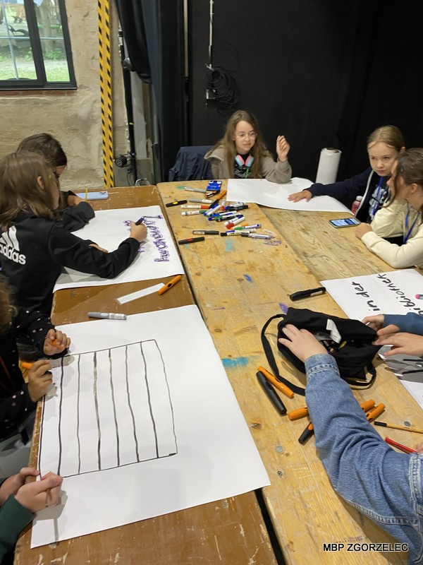 Zdjęcie przedstawia grupę dzieci siedzących przy drewnianym stole i biorących udział w zajęciach plastycznych. Na stole znajdują się kolorowy papier, markery, kleje, taśmy i inne materiały dekoracyjne. Dzieci pracują nad plakatami.