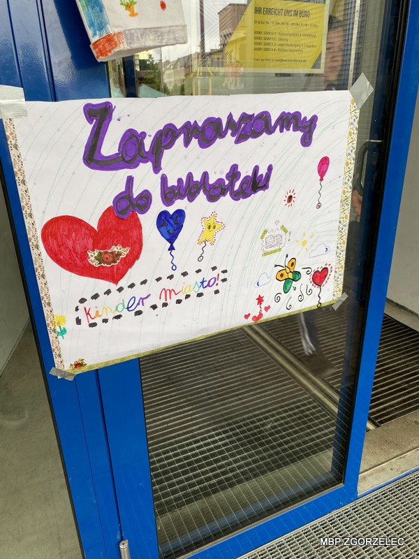 Zdjęcie przedstawia ręcznie wykonany, kolorowy plakat przyklejony do niebieskich drzwi. Na plakacie widnieje napis 'Zapraszamy do biblioteki' dużymi fioletowymi literami oraz 'Kinder miasto!' w mniejszych, wielokolorowych literach. Plakat ozdobiony jest rysunkami serc, balonów, gwiazdek i innymi dekoracyjnymi elementami.