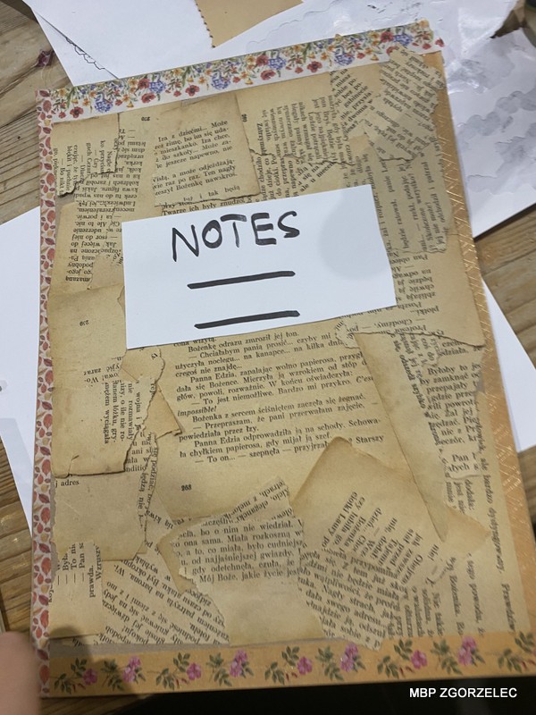 Notes z okładką z porwanych stron książek, ozdobiony kwiatową taśmą i etykietą z napisem „NOTES”.