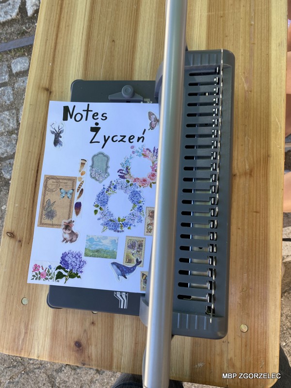 Zdjęcie przedstawia drewniany stół, na którym znajduje się metalowa bindownica oraz ozdobiona okładka zeszytu z napisem 'Notes Życzeń'. Okładka udekorowana jest naklejkami z motywami kwiatów, zwierząt i innymi elementami ozdobnymi.