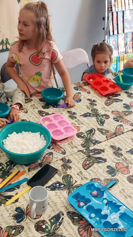 Grupa dzieci siedzących wokół stołu z obrusem w motyle. Na stole znajdują się kolorowe silikonowe foremki, plastikowe miski, duża miska z mydłem glicerynowym w kostkach, plastikowe łyżki, kubek oraz smartfon. Dzieci uczestniczą w aktywności związanej z wykorzystaniem foremek i materiału z miski.