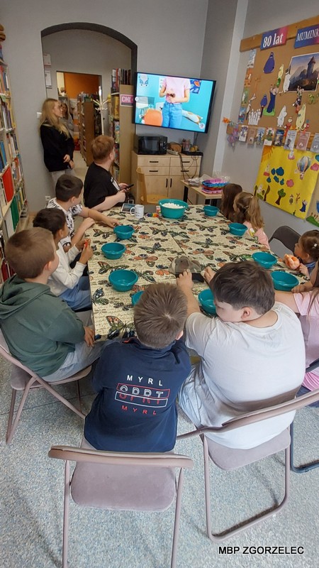 Dzieci i dorośli oglądają na telewizorze materiał edukacyjny przy stole z zielonymi miskami
