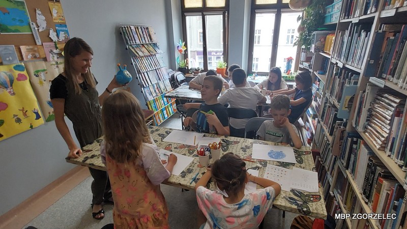 Zdjęcie w pomieszczeniu biblioteki przedstawia grupę dzieci oraz bibliotekarkę trzymającą maskotkę. Dzieci siedzą wokół stołu i uczestniczą w zajęciach rysunkowych lub pisemnych. Po prawej stronie znajdują się regały z książkami, a po lewej plakaty na ścianie.