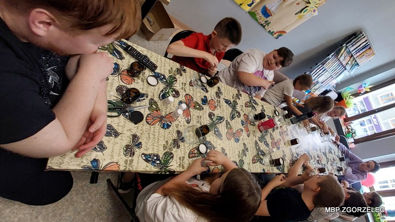 Dzieci pracują przy stole z małymi słoiczkami w udekorowanej bibliotece podczas warsztatów tworzenia świec sojowych.